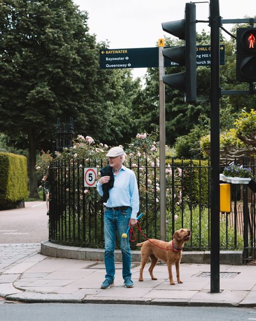 A man standing at a pedestrian crossing on a city street, holding a black bag over his shoulder and accompanied by a brown Labrador retriever on a red leash. The man is wearing a light grey cap, a lig